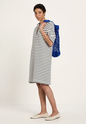 Frau in weißem knielangem Kleid mit schwarzen Streifen, die eine blaue Netztasche über der Schulter trägt, beige Ballerinas an den Füßen, vor einfachem Hintergrund.