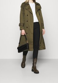 Olivově zelený trenčkot s dvouřadým zapínáním, hnědými knoflíky a manžetami na pásek, kombinovaný s černými kalhotami a hnědými kotníkovými botami. Černá kabelka.