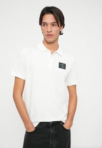 Polo alb din bumbac cu guler clasic și mâneci scurte, având o pată rectangulată neagră pe piept cu text. Purtați cu blugi închiși la culoare.