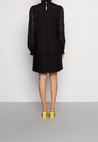 Vestido negro de encaje con mangas largas, cuello alto y detalle de abertura en la parte trasera. Combinado con sandalias amarillas de tacón bloque. Fondo gris liso.