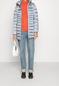 Persona con una chaqueta de plumas gris claro sobre un suéter rojo de canalé, jeans azules con dobladillo, botas negras hasta el tobillo, sosteniendo un bolso blanco al lado.