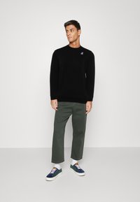 Felpa nera con collo a giro e un piccolo logo ricamato, abbinata a pantaloni larghi verde oliva e scarpe da ginnastica multicolori.