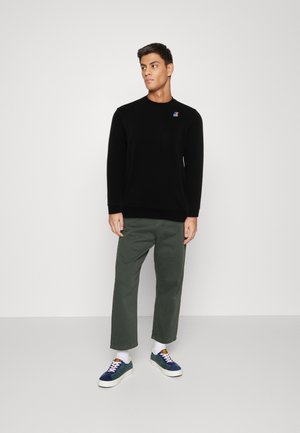 Felpa nera con collo a giro e un piccolo logo ricamato, abbinata a pantaloni larghi verde oliva e scarpe da ginnastica multicolori.