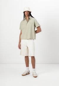 Camicia a maniche corte verde chiaro con due tasche sul petto, abbinata a pantaloni corti beige fino al ginocchio e sneakers beige con dettagli scuri.