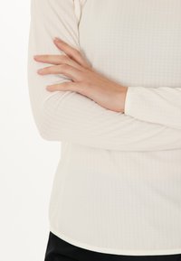 Helle, langärmlige Oberteil mit einem strukturierten Muster, das einen Rundhalsausschnitt und ergänzende Abschlüsse aufweist. Hand auf dem Ärmel positioniert.