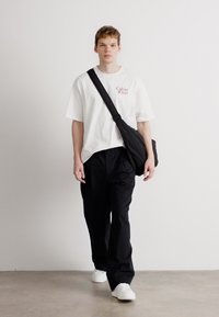 Tricou alb din bumbac cu logo-ul roșu "Calvin Klein", asociat cu pantaloni negri largi și adidași albi, purtând o geantă de umăr neagră.