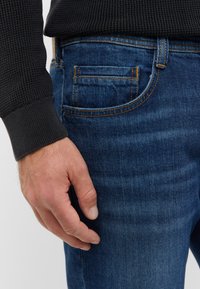 Jeans in denim blu con cuciture a contrasto, una tasca posteriore e un design aderente. Mano appoggiata sulla tasca, con un maglione nero texturizzato visibile.