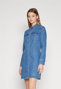 Rochie din denim cu mâneci lungi, culoare albastru deschis, două buzunare pe piept, închidere cu nasturi în față și detalii de cusătură albă.