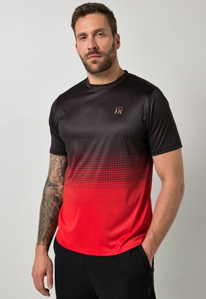 Mężczyzna z brodą i tatuażem na prawej ręce, ubrany w czarno-czerwoną koszulkę z krótkim rękawem w gradient i czarne spodnie, stoi z lewą ręką w kieszeni.