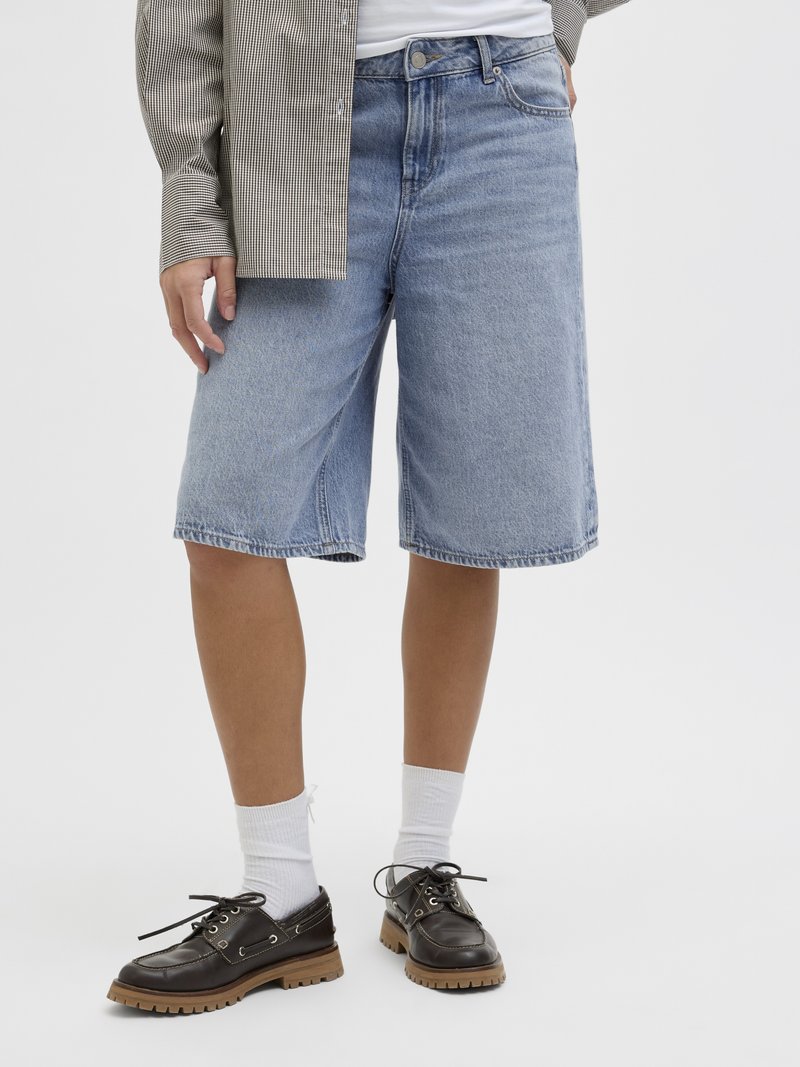 Shorts de ganga azul claro com um corte relaxado, dois bolsos frontais e uma textura suave, combinados com sapatos pretos com atacadores e meias brancas.