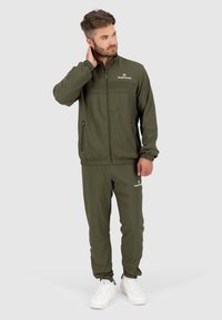 Olijfgroen trainingspak met elastische boorden en zoom, uitgerust met een ritsjack en een broek met zijzakken, gecombineerd met witte sneakers.
