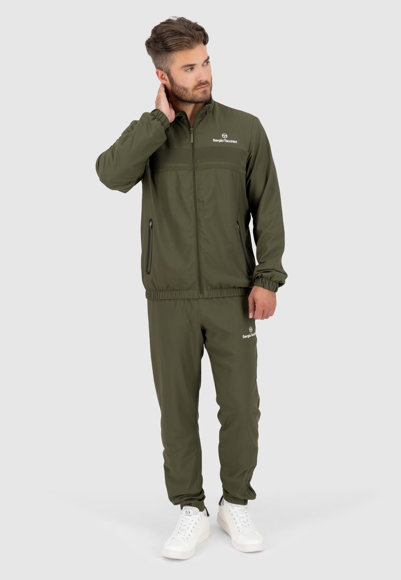 Olijfgroen trainingspak met elastische boorden en zoom, uitgerust met een ritsjack en een broek met zijzakken, gecombineerd met witte sneakers.