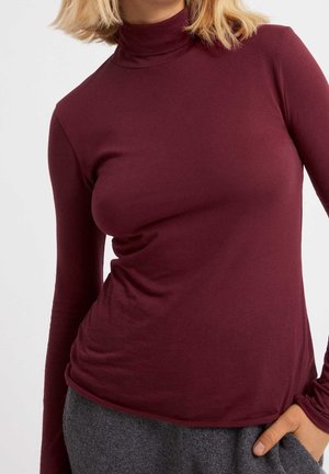 Kobieta w dopasowanym bordowym golfie z długim rękawem i szarych spodniach, z prawą ręką włożoną do kieszeni.