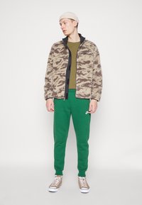 Tan patterned fleece jacket with black accents, olive green shirt, green sweatpants, and tan sneakers on a standing figure.