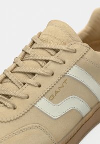 Baskets en daim beige avec lacets beiges et accents en cuir blanc. Comprennent un logo de marque doré embossé, une semelle en caoutchouc texturée et des détails cousus.