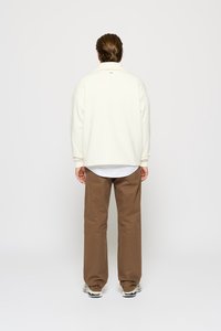 Felpa color crema con colletto ripiegato, abbinata a pantaloni marroni comodi e larghi. Canottiera bianca visibile all'orlo inferiore. Calzature casual.