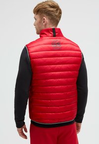 Rote gepolsterte Weste mit hohem Kragen, ausgestattet mit einem schwarzen Reißverschluss und einem Logodetail auf der Rückseite. Die Ärmel sind schwarz und der Saum hat einen kontrastierenden Streifen.