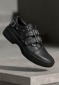 Black leather sneaker with textured upper, multiple buckle straps, and a chunky sole. Features silver-tone hardware and a smooth finish.