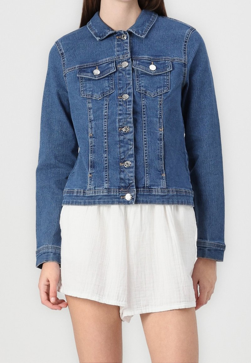 Femme portant une veste en jean bleu ajustée avec des boutons pression et deux poches poitrine, sur un short blanc texturé.
