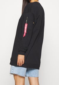 Sweat-shirt noir à manches longues, avec une poche zippée sur le bras et une étiquette rouge. Porté avec un short en denim bleu clair.