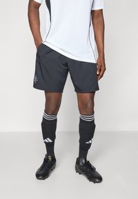 Sorte atletiske shorts med et logo, parret med knæhøje sorte sokker med hvide striber og sorte fodboldstøvler. Hvid skjorte med sorte detaljer.