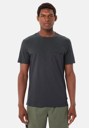 Homme portant un t-shirt à manches courtes gris foncé et un pantalon vert olive, debout avec les bras relâchés contre un fond blanc uni.