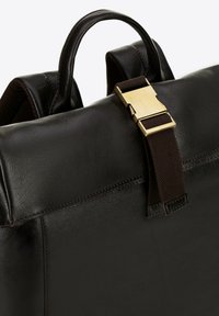 Hnědý kožený batoh s texturovaným povrchem, který má zlatou sponu a hnědý textilní popruh. Zdůrazňuje elegantní design a praktické kování.