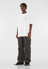 White oversized t-shirt with red print on the chest paired with black cargo pants featuring multiple pockets and a loose fit. Black sneakers.