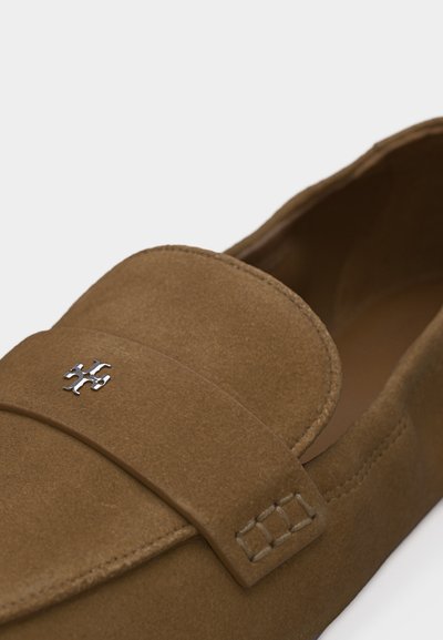 Καφέ μοκασίνι από σουέντ με μικρό ασημί διακοσμητικό λογότυπο στη λουρίδα πάνω από το παπούτσι.
