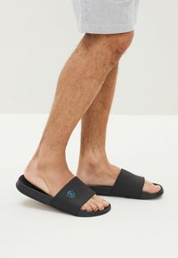 Svarta halksandaler med en bred, texturerad rem, öppen tådesign och en blå grafisk detalj på remmen. Platt sula för komfort.