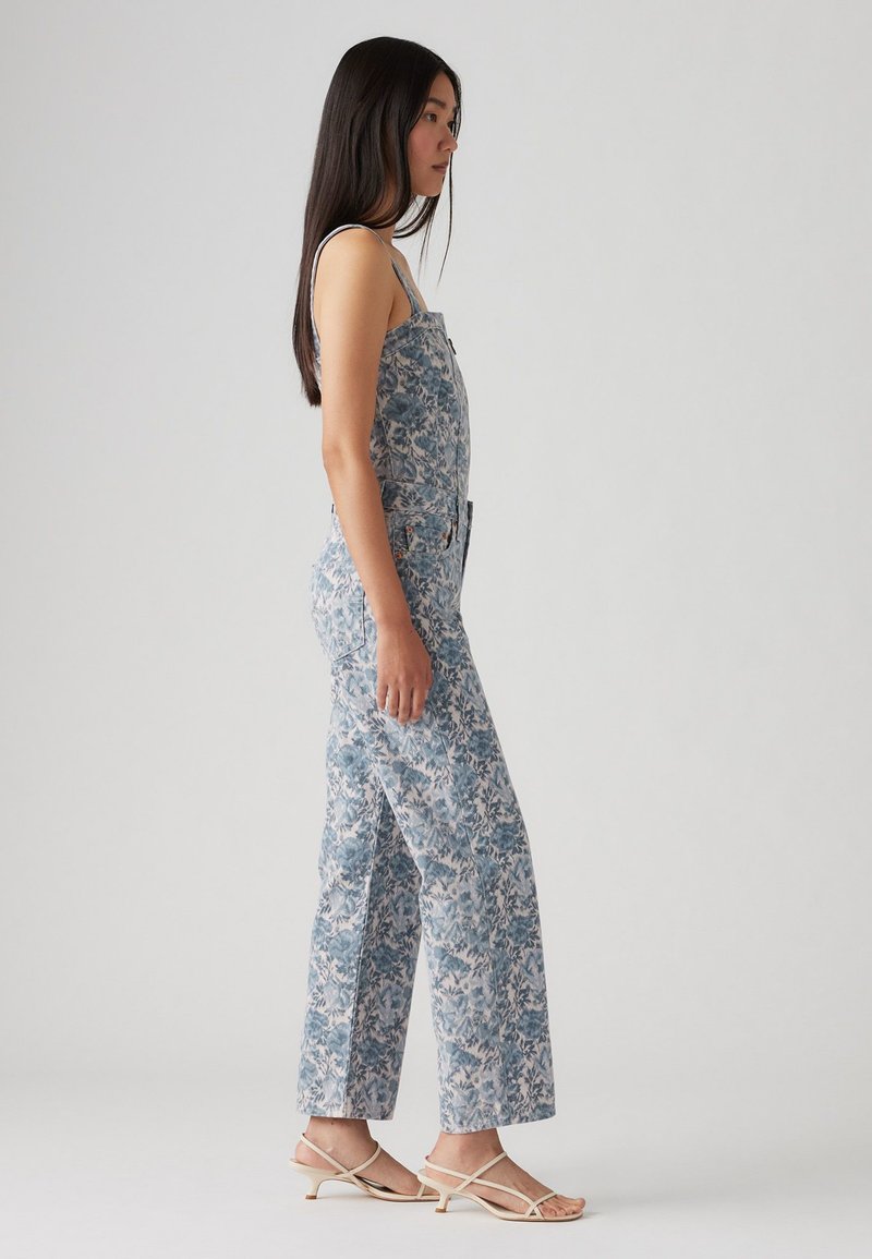 Jumpsuit à motifs floraux en bleu clair et blanc, avec un bas à jambes larges et un buste ajusté. Des sandales à brides étroites complètent la tenue.