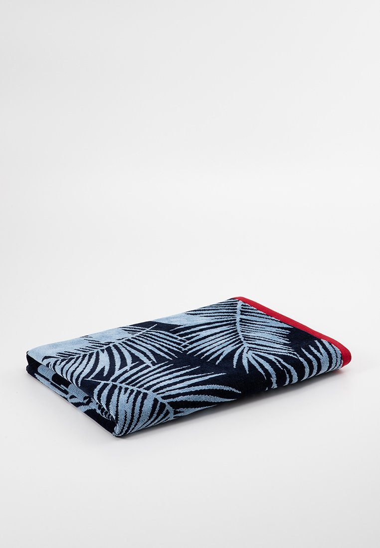 Toalla de playa rectangular en azul y marino con un patrón de hojas de palma. Presenta un borde rojo a lo largo de un lado. Material de algodón suave y texturizado.