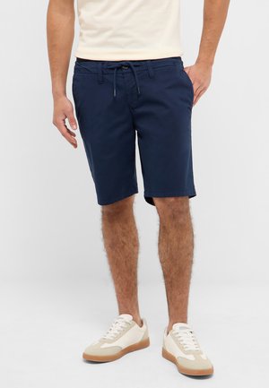 Mann trägt marineblaue Shorts mit Kordelzug, cremefarbene Sneaker und ein hellbeiges T-Shirt, steht mit entspannt an den Seiten hängenden Händen.