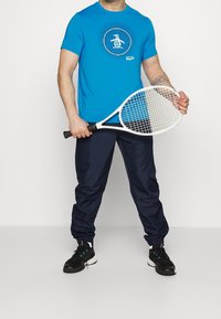 Man som bär en blå t-shirt med pingvinlogo, marinblå joggerbyxor och svarta sneakers håller en vit tennisracket mot en vit bakgrund.