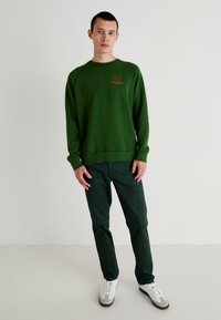 Grønn genser med rund hals, ribbestrikkede mansjetter og nederkant, med en oransje logo. Kombinert med mørkegrønne bukser og hvite joggesko.