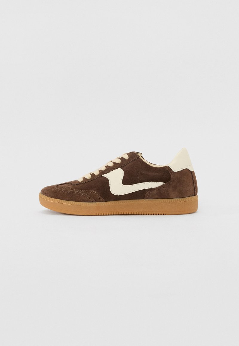 Sneaker en suedine marron avec des accents crème et une semelle en caoutchouc gomme, présentant un logo distinctif en forme de swoosh sur le côté.