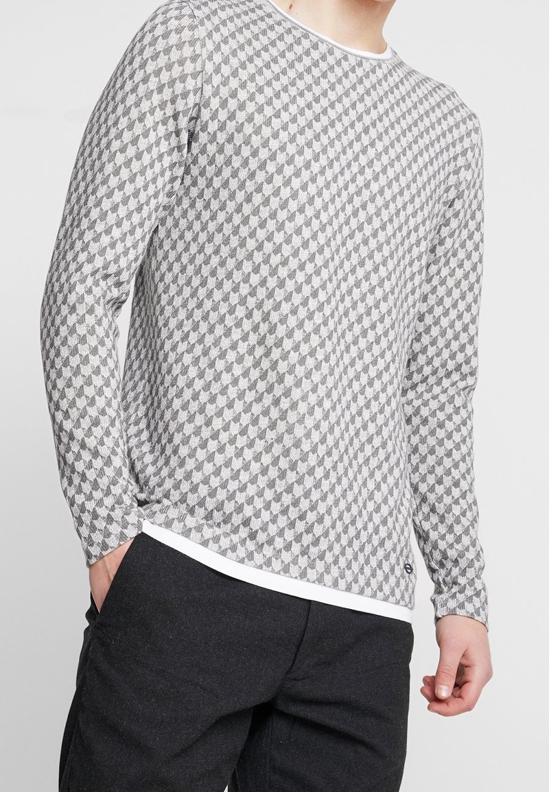 Homme portant un pull à manches longues gris et blanc à motifs, pantalon noir, une main dans la poche, sur un fond uni.