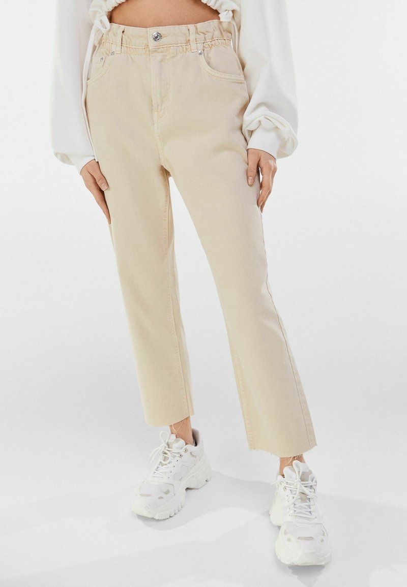Pantalon court beige taille haute avec ourlet brut, porté avec des baskets blanches épaisses et un haut blanc à manches longues avec des poignets élastiques.