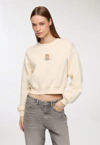 Felpa corta crema con maniche lunghe, caratterizzata da una stampa di un orso dei cartoni e un design testuale. Abbinata a jeans grigi a vita alta, che mostrano un tessuto testurizzato.