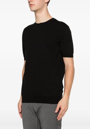 Uomo che indossa un maglione nero semplice a maniche corte e pantaloni grigi, in piedi su uno sfondo bianco, volto leggermente a sinistra.