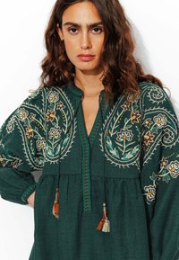 Tunique brodée vert foncé avec des motifs floraux, décolleté en V, détails à franges et manches bouffantes. Fabriquée en tissu texturé.