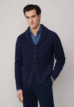 Cardigan en maille côtelée marine avec col châle, doté d'une fermeture à boutons, de deux poches avant et d'un revers contrastant aux poignets. Porté par-dessus une chemise bleue.