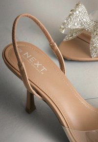 Naakte sandalen met hoge hak, voorzien van een doorzichtige band en een zachte, suède-achtige enkelband. Decoratieve strik met steentjes op een aangrenzende schoen.