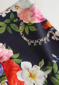 Kvetinová látka s tmavým pozadím, veľké ružové, biele a červené kvety. Obsahuje vzor pripomínajúci reťaz s nápisom „BAKER“ v zlatej farbe.