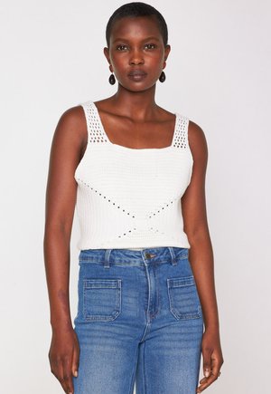 Donna che indossa un top a maniche corte bianco in maglia con scollatura quadrata e jeans blu a vita alta, in piedi contro uno sfondo chiaro e uniforme.