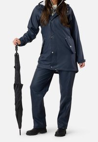Chaqueta de lluvia impermeable en color navy con bolsillos, combinada con pantalones a juego y botas de goma negras, sosteniendo un paraguas negro. Textura suave en toda la prenda.