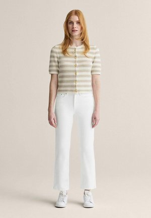 Kvinde med rødt hår iført kortærmet beige og hvidstribet knappet top, hvide bukser med lige ben og hvide sneakers, stående på ensfarvet baggrund.