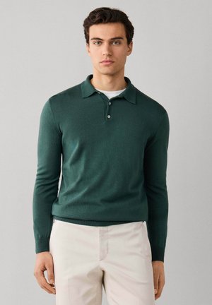 Joven con polo de manga larga verde oscuro y pantalones beige claros, de pie frente a un fondo gris liso.