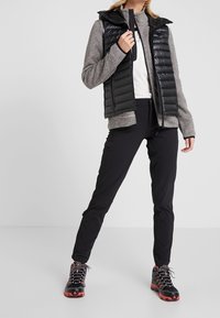 Chaleco acolchado negro sobre un suéter gris texturizado con cremallera, combinado con pantalones negros y botas de senderismo. El conjunto presenta materiales en capas y un diseño deportivo.