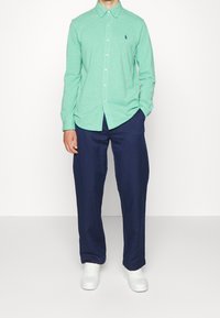 Man wearing long-sleeve mint green button-up shirt, navy blue pants, white sneakers, standing with one hand in pocket against plain background.